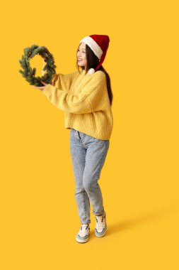 Sarı arka planda Noel çelengi olan Noel Baba şapkalı mutlu genç Asyalı kadın.