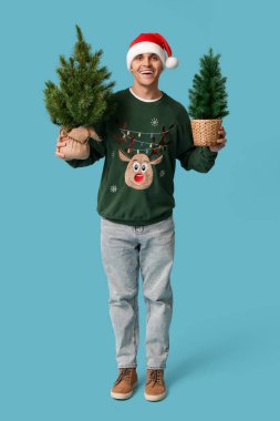 Mavi arka planda Noel ağaçları olan Noel Baba şapkalı genç adam.