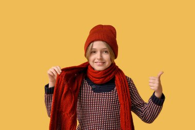 Sarı arka planda baş parmak hareketi gösteren kışlık elbiseli şirin bir çocuk.