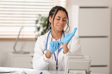 Genç Afro-Amerikalı kadın doktor klinikteki masada tıbbi eldiven takıyor.