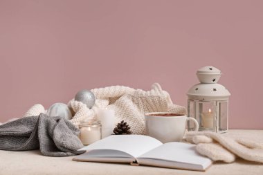 Kitap, bir fincan kakao, Noel süslemeleri ve pembe duvarın yanındaki grunge beyaz masa üzerindeki kalın giysilerle güzel bir kış kompozisyonu.