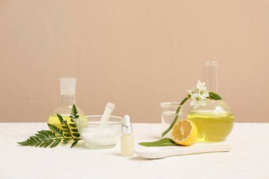 Bej duvarın yanındaki beyaz masada doğal kozmetik ürünleri hazırlamak için malzemeler ve numunelerle dolu laboratuvar vitrinleri.