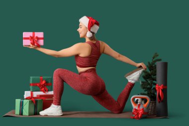 Noel hediyesi kutuları, kettlebell ve yoga minderiyle yeşil arka planda spor yapan genç bir kadın.