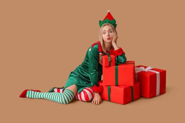 Kahverengi arka planda Noel hediyesi kutuları olan elf kostümlü düşünceli genç bir kadın.