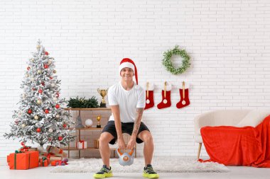 Noel Baba şapkalı genç adam Noel için dekore edilmiş oturma odasında kettlebell ile antrenman yapıyor.