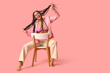 Saçları örgülü, pembe arka planda oturan seksi Afro-Amerikan kadın.