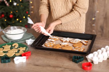 Genç bir kadın Noel arifesinde mutfaktaki masada zencefilli kurabiye süslüyor.
