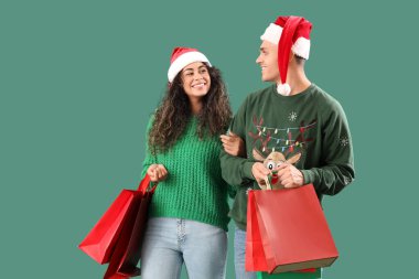 Yeşil arka planda alışveriş torbaları olan Noel Baba şapkalı mutlu bir çift.