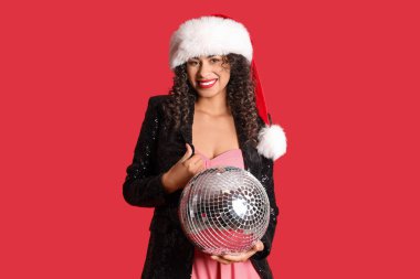 Kırmızı arka planda disko topu olan Noel Baba şapkalı güzel bir Afro-Amerikalı kadın. Yeni yıl kutlaması