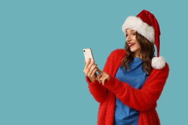 Noel Baba şapkalı genç kadın mavi arka planda selfie çekiyor.