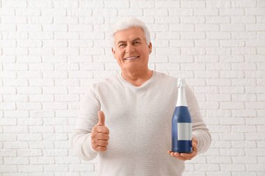 Bir şişe şampanyayla Noel 'i kutlayan ve beyaz tuğladan arka planda baş parmak hareketi yapan kıdemli bir adam.