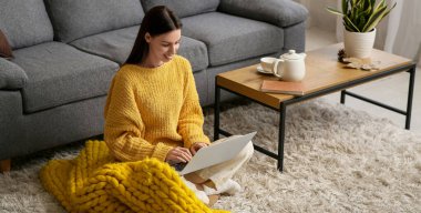 Sıcak sarı ekoseli ve dizüstü bilgisayarlı genç bir kadın oturma odasında yerde oturuyor.