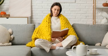 Oturma odasındaki kanepede sıcak sarı ekoseli kitap okuyan genç bir kadın.