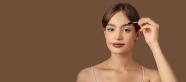 Kahverengi arka planda kaşlarının şeklini düzelten genç bir kadın.