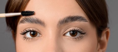 Gri arka planda kaşlarını düzelten genç bir kadın, yakın plan.