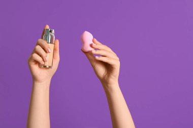 Makyaj süngeri ve mor arka planda bir şişe fondöten olan kadın elleri.