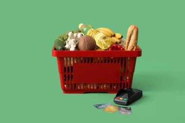 Ödeme terminali ve kredi kartlarıyla dolu alışveriş sepeti yeşil arka planda.