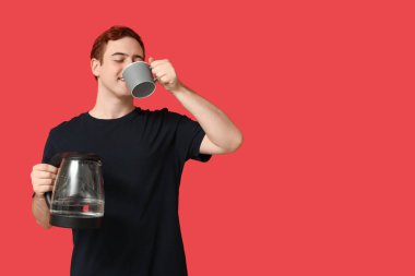 Modern elektrikli çaydanlığı olan yakışıklı genç adam kırmızı arka planda kahve içiyor.