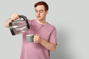 Yakışıklı genç adam, modern elektrikli çaydanlıktan gri arka planda kupaya sıcak su döküyor.