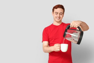 Yakışıklı genç adam modern elektrikli çaydanlıktan gri arka planda fincana sıcak su dolduruyor.