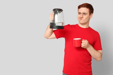 Modern elektrikli su ısıtıcısı ve gri arka planda kupası olan düşünceli genç bir adam.