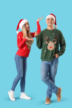 Mavi arka planda Noel Baba şapkalı ve Noel kazaklı genç bir çift.