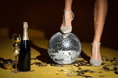 Şampanyalı genç kadın bacakları, disko topu ve sarı arka planda parti dekoru. Yeni yıl kutlaması
