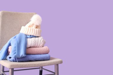Stacked sweaters with hat on chair against lilac background
