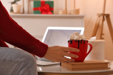 Bir kadın masadan laptopuyla sıcak çikolata alıyor, şenlikli oturma odasında, yakın plan.