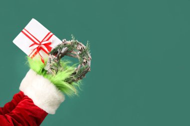 Hairy hands of creature in Santa costume with Christmas gift box and wreath on green background clipart