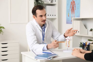 Male doctor explaining spinal anatomy with vertebral column model to patient in clinic clipart