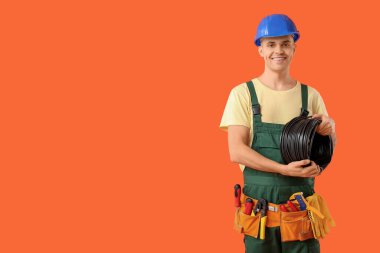 Aletleri, kaskı ve kablosu turuncu arka planda olan genç bir elektrikçi.