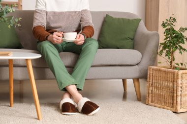 Terlikli bir adam oturma odasında bir fincan kahveyle kanepede oturuyor.