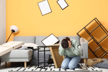 Depremden sonra dağınık mobilyalarla oturma odasında oturan genç bir kadın.