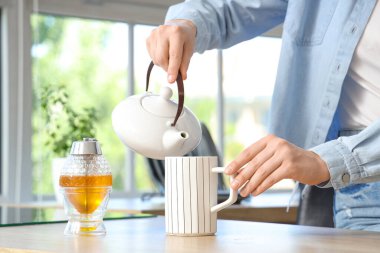 Genç bir kadın mutfaktaki çaydanlıktan fincana içecek dolduruyor.