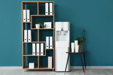 Masasında kağıt bardaklar ve mavi duvarın yanında raf ünitesi olan modern su soğutucusu.