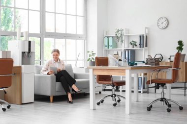 Ofisteki su soğutucusunun yanındaki koltukta oturan tablet bilgisayarlı iş kadını.