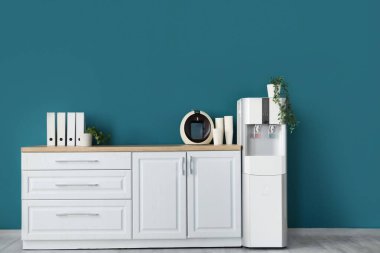 Tezgahta kağıt bardaklı kahve makinesi ve ofis mutfağında mavi duvarın yanında su soğutucusu.