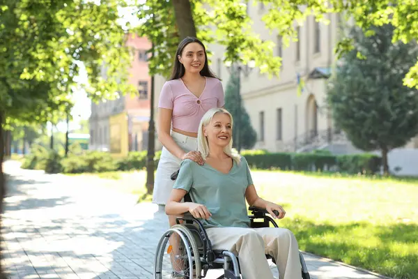 Tekerlekli sandalyedeki yaşlı kadın ve kızı dışarıda birlikte vakit geçiriyorlar.