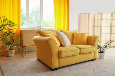 Rahat bir kanepe, ev bitkisi ve oturma odasının içinde sarı perdeler.