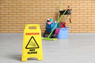 Temizlik malzemeleri, uyarı levhası ve tuğla duvarın yanındaki zemin paspası olan kapıcı vagonu.