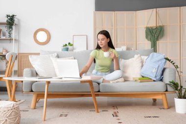 Evde dizüstü bilgisayar kullanan bir fincan kahveli kız öğrenci.