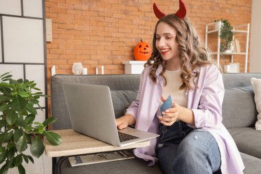 Şeytan boynuzlu, kredi kartı ve dizüstü bilgisayarlı genç bir kadın Cadılar Bayramı için evdeki kanepede alışveriş yapıyor.
