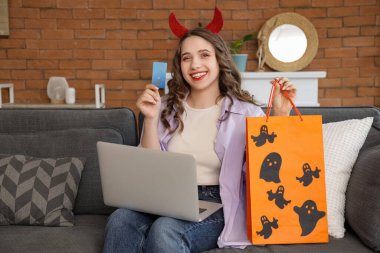 Şeytan boynuzlu, kredi kartı ve dizüstü bilgisayarlı genç bir kadın Cadılar Bayramı için evdeki kanepede alışveriş yapıyor.