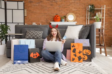 Şeytan boynuzlu genç bir kadın kredi kartı ve dizüstü bilgisayarla Cadılar Bayramı için internette alışveriş yapıyor.