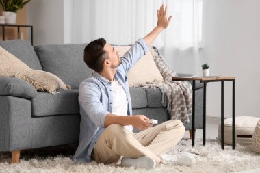 Klima kumandalı genç adam bej duvarın yanında yerde oturuyor.