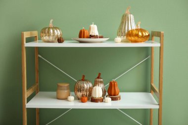 Yeşil duvarın yanında cam balkabağı, tarçın ve balkabağı şeklinde mumlar olan raf ünitesi.