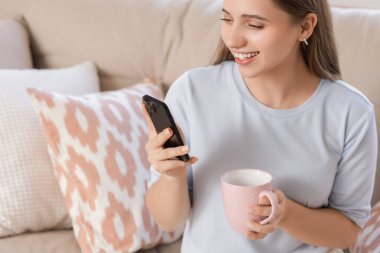 Evdeki kanepede çay fincanı ile gülümseyen genç kadın, yakın plan.