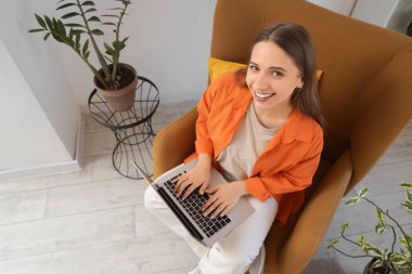 Evdeki koltukta dizüstü bilgisayar kullanan gülümseyen genç kadın.