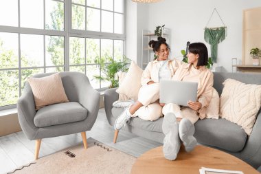 Evdeki kanepede dizüstü bilgisayar kullanan genç lezbiyen çift.